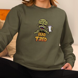 K&ouml;nnte beinhalten: Olivgr&uuml;nes Sweatshirt mit einem Cartoon-Zombie, der eine Kaffeetasse h&auml;lt. Der Zombie hat einen m&uuml;den Gesichtsausdruck, und der Text "DEAD TIRED" ist orange und gelb. Das Sweatshirt hat einen Rundhalsausschnitt.
