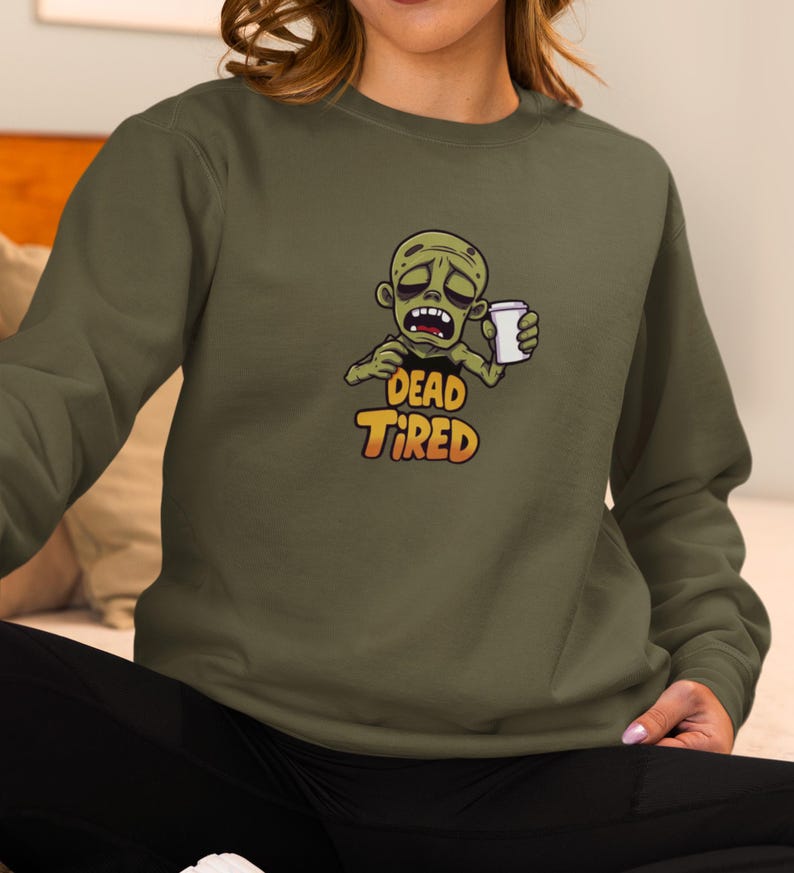K&ouml;nnte beinhalten: Olivgr&uuml;nes Sweatshirt mit einem Cartoon-Zombie, der eine Kaffeetasse h&auml;lt. Der Zombie hat einen m&uuml;den Gesichtsausdruck, und der Text "DEAD TIRED" ist orange und gelb. Das Sweatshirt hat einen Rundhalsausschnitt.