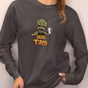 K&ouml;nnte beinhalten: Dunkelgraues Langarmshirt mit einem Cartoon-Zombie, der eine Kaffeetasse h&auml;lt. Der Zombie hat einen m&uuml;den Gesichtsausdruck, und der Text "DEAD TIRED" steht darunter in Orange und Gelb.