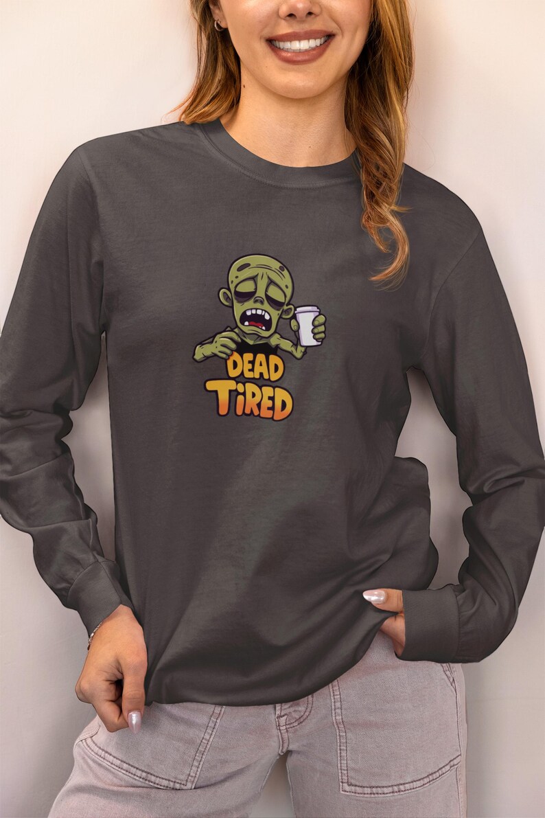 K&ouml;nnte beinhalten: Dunkelgraues Langarmshirt mit einem Cartoon-Zombie, der eine Kaffeetasse h&auml;lt. Der Zombie hat einen m&uuml;den Gesichtsausdruck, und der Text "DEAD TIRED" steht darunter in Orange und Gelb.