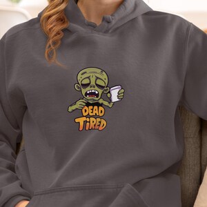 K&ouml;nnte beinhalten: Dunkelgrauer Kapuzenpullover mit einem Cartoon-Zombie, der eine Kaffeetasse h&auml;lt. Der Zombie ist gr&uuml;n und hat einen m&uuml;den Gesichtsausdruck. Die Worte "DEAD TIRED" sind darunter in Orange und Gelb gedruckt.