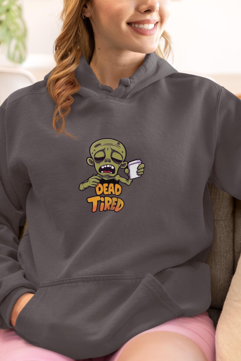 K&ouml;nnte beinhalten: Dunkelgrauer Kapuzenpullover mit einem Cartoon-Zombie, der eine Kaffeetasse h&auml;lt. Der Zombie ist gr&uuml;n und hat einen m&uuml;den Gesichtsausdruck. Die Worte "DEAD TIRED" sind darunter in Orange und Gelb gedruckt.