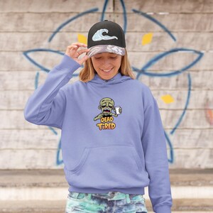 K&ouml;nnte beinhalten: Ein hellvioletter Kapuzenpullover mit einer Vordertasche und einer Cartoon-Zombie-Grafik, die eine Kaffeetasse h&auml;lt. Der Text "DEAD TIRED" steht unter dem Zombie. Die Person tr&auml;gt eine schwarze Baseballkappe mit einem Wellenmuster.