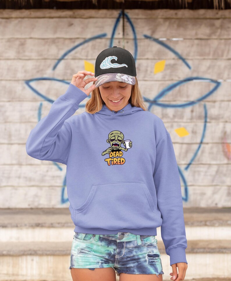 K&ouml;nnte beinhalten: Ein hellvioletter Kapuzenpullover mit einer Vordertasche und einer Cartoon-Zombie-Grafik, die eine Kaffeetasse h&auml;lt. Der Text "DEAD TIRED" steht unter dem Zombie. Die Person tr&auml;gt eine schwarze Baseballkappe mit einem Wellenmuster.
