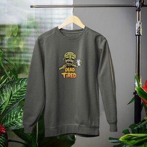 K&ouml;nnte beinhalten: Dunkelgraues Sweatshirt mit einem Cartoon-Zombie, der eine Kaffeetasse h&auml;lt. Der Zombie hat einen m&uuml;den Gesichtsausdruck, und der Text "DEAD TIRED" ist in Orange und Gelb unter dem Bild aufgedruckt. Das Sweatshirt wird auf einem Holzb&uuml;gel pr&auml;sentiert.