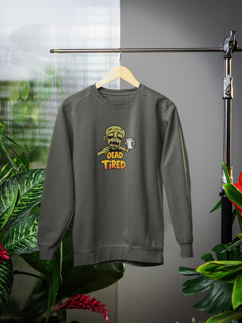 K&ouml;nnte beinhalten: Dunkelgraues Sweatshirt mit einem Cartoon-Zombie, der eine Kaffeetasse h&auml;lt. Der Zombie hat einen m&uuml;den Gesichtsausdruck, und der Text "DEAD TIRED" ist in Orange und Gelb unter dem Bild aufgedruckt. Das Sweatshirt wird auf einem Holzb&uuml;gel pr&auml;sentiert.