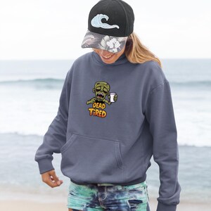 K&ouml;nnte beinhalten: Ein marineblauer Kapuzenpullover mit einer Vordertasche und einer Grafik eines Zombies, der eine Kaffeetasse h&auml;lt, mit den Worten "DEAD TIRED". Die Person tr&auml;gt eine schwarze Baseballkappe mit einem Wellenmuster.