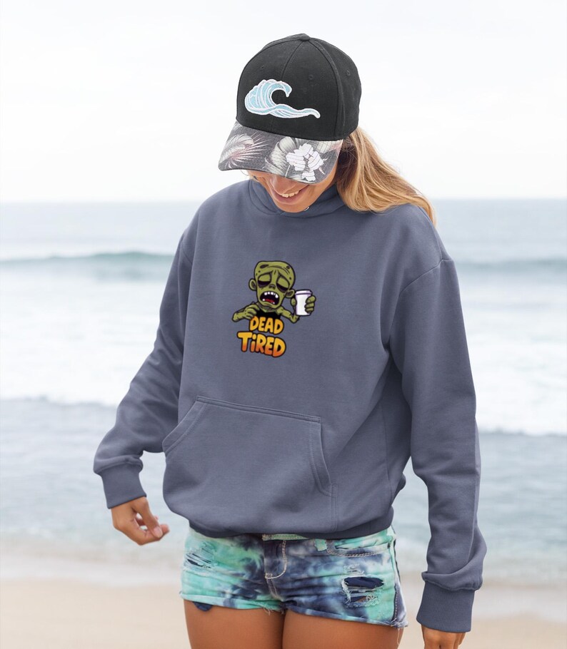K&ouml;nnte beinhalten: Ein marineblauer Kapuzenpullover mit einer Vordertasche und einer Grafik eines Zombies, der eine Kaffeetasse h&auml;lt, mit den Worten "DEAD TIRED". Die Person tr&auml;gt eine schwarze Baseballkappe mit einem Wellenmuster.