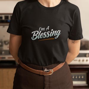 May include: Black crew neck t-shirt with the text "I'm A Blessing" in a stylized font. The shirt is paired with brown trousers and a brown belt with a gold buckle. The overall style is casual and comfortable.