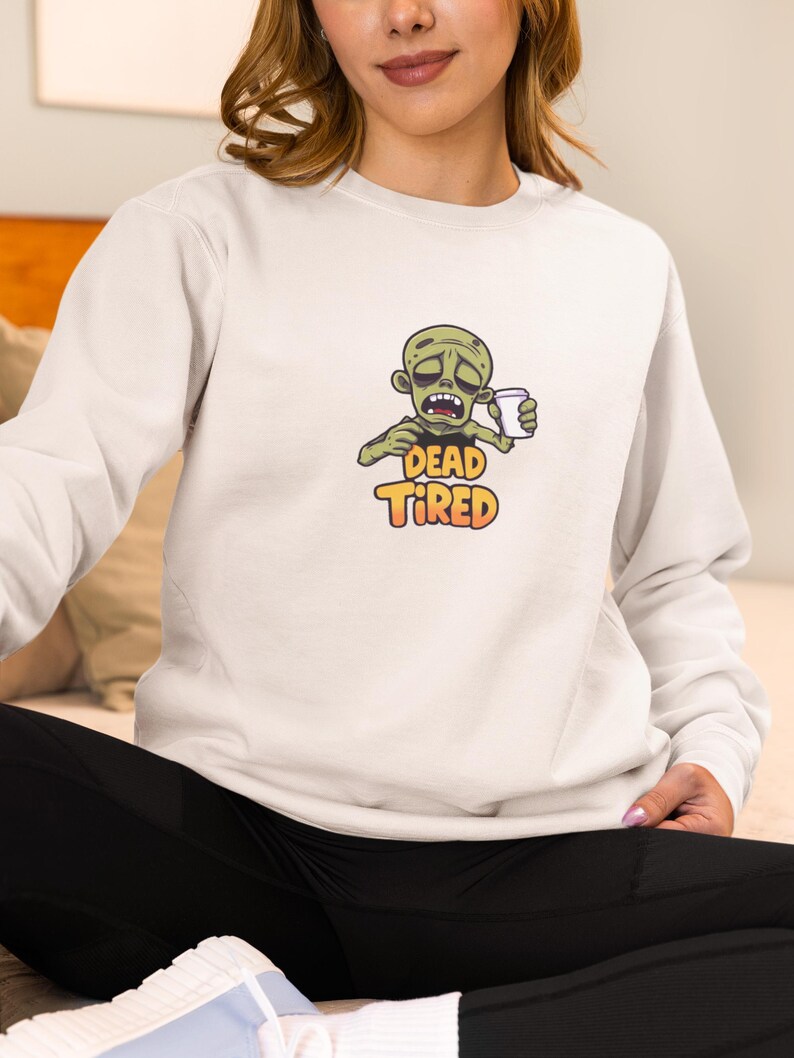K&ouml;nnte beinhalten: Hellgraues Sweatshirt mit einem Cartoon-Zombie, der eine Kaffeetasse h&auml;lt. Der Zombie hat gr&uuml;ne Haut und einen m&uuml;den Gesichtsausdruck. Die Worte "DEAD TIRED" sind darunter in Orange und Gelb gedruckt.