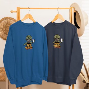 K&ouml;nnte beinhalten: Zwei Sweatshirts mit Rundhalsausschnitt, eines in Dunkelt&uuml;rkis und das andere in Marineblau, jeweils mit einem Zombie-Motiv, das eine Kaffeetasse h&auml;lt. Der Text "DEAD TIRED" steht darunter. Die Sweatshirts h&auml;ngen an einem Holzst&auml;nder.