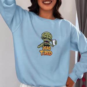 K&ouml;nnte beinhalten: Hellblaues Sweatshirt mit einem Cartoon-Zombie, der eine Kaffeetasse h&auml;lt. Der Zombie hat einen m&uuml;den Gesichtsausdruck, und der Text "DEAD TIRED" ist in Orange und Gelb dargestellt. Das Sweatshirt hat lange &Auml;rmel und einen Rundhalsausschnitt.