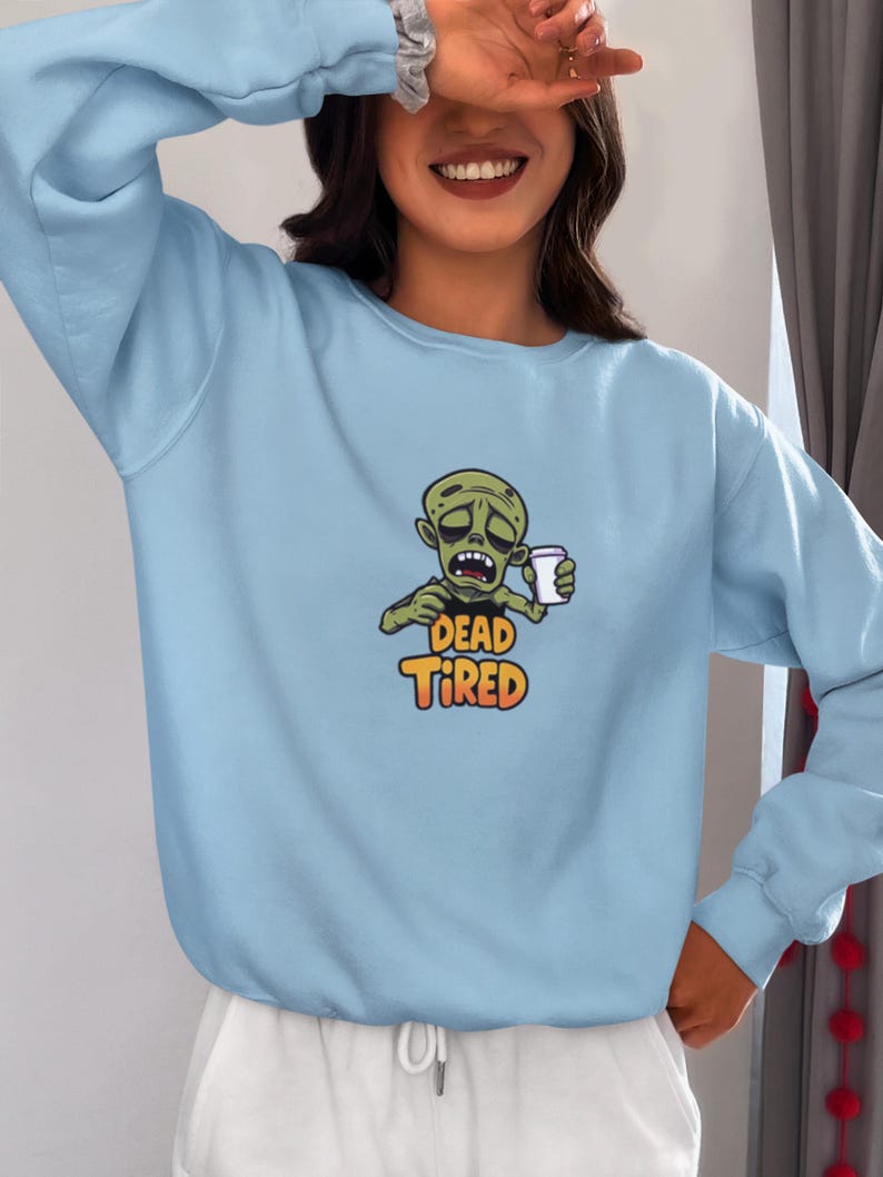K&ouml;nnte beinhalten: Hellblaues Sweatshirt mit einem Cartoon-Zombie, der eine Kaffeetasse h&auml;lt. Der Zombie hat einen m&uuml;den Gesichtsausdruck, und der Text "DEAD TIRED" ist in Orange und Gelb dargestellt. Das Sweatshirt hat lange &Auml;rmel und einen Rundhalsausschnitt.