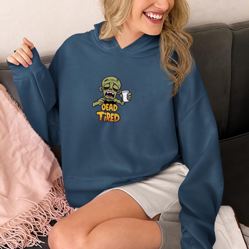 K&ouml;nnte beinhalten: Marineblauer Hoodie mit einem Cartoon-Zombie, der eine Kaffeetasse h&auml;lt. Unter der Abbildung des Zombies steht der Text &bdquo;DEAD TIRED&ldquo;. Der Hoodie hat eine Kapuze und eine Vordertasche.