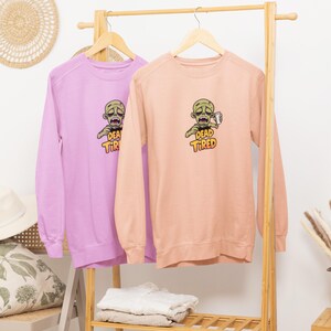 K&ouml;nnte beinhalten: Zwei lang&auml;rmelige Sweatshirts, eines in Lavendel und eines in Pfirsich, h&auml;ngen an einem Holzgestell. Jedes Sweatshirt zeigt eine Grafik eines Zombies, der eine Tasse h&auml;lt, mit den Worten "DEAD TIRED" in Gelb und Orange.