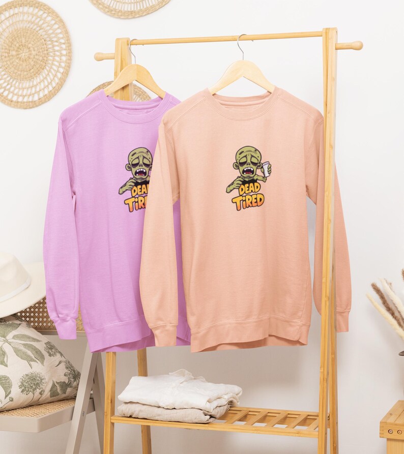 K&ouml;nnte beinhalten: Zwei lang&auml;rmelige Sweatshirts, eines in Lavendel und eines in Pfirsich, h&auml;ngen an einem Holzgestell. Jedes Sweatshirt zeigt eine Grafik eines Zombies, der eine Tasse h&auml;lt, mit den Worten "DEAD TIRED" in Gelb und Orange.