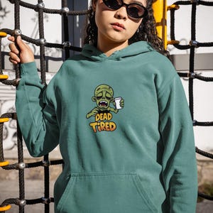 K&ouml;nnte beinhalten: T&uuml;rkisfarbener Kapuzenpullover mit einer Vordertasche und einer Grafik eines Zombies, der eine Kaffeetasse h&auml;lt. Der Text "DEAD TIRED" befindet sich unter der Zombie-Illustration. Die Person auf dem Bild tr&auml;gt eine Sonnenbrille und hat ihr Haar in zwei Dutt.