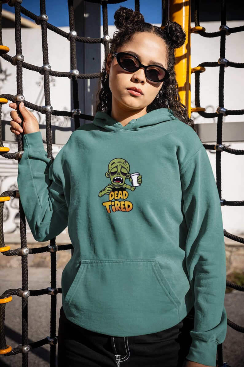 K&ouml;nnte beinhalten: T&uuml;rkisfarbener Kapuzenpullover mit einer Vordertasche und einer Grafik eines Zombies, der eine Kaffeetasse h&auml;lt. Der Text "DEAD TIRED" befindet sich unter der Zombie-Illustration. Die Person auf dem Bild tr&auml;gt eine Sonnenbrille und hat ihr Haar in zwei Dutt.