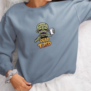 K&ouml;nnte beinhalten: Hellblaues Sweatshirt mit einem Cartoon-Zombie, der eine Kaffeetasse h&auml;lt. Der Zombie hat einen m&uuml;den Gesichtsausdruck, und die Worte "DEAD TIRED" sind darunter in Orange und Gelb aufgedruckt. Das Sweatshirt ist einfarbig.