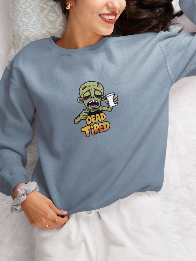 K&ouml;nnte beinhalten: Hellblaues Sweatshirt mit einem Cartoon-Zombie, der eine Kaffeetasse h&auml;lt. Der Zombie hat einen m&uuml;den Gesichtsausdruck, und die Worte "DEAD TIRED" sind darunter in Orange und Gelb aufgedruckt. Das Sweatshirt ist einfarbig.