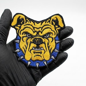 Könnte beinhalten: Gestickter Aufnäher mit dem Kopf einer Bulldogge mit grimmigem Gesichtsausdruck. Der Aufnäher hat ein gelb-schwarzes Farbschema mit einem blauen Kragen mit silbernen Stacheln. Der Aufnäher wird gegen einen schwarzen Handschuh gehalten.