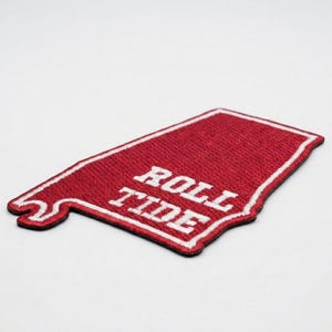 May include: A red and white embroidered patch in the shape of the state of Alabama. The patch features the words "ROLL TIDE" in white block letters. The patch has a white border and a black edge.