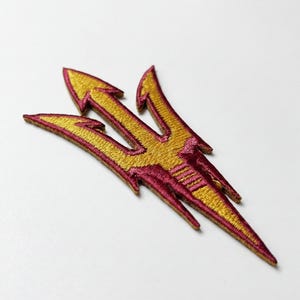 Pode incluir: Patch bordado com um design de tridente dourado com detalhes em bordô. O tridente tem três pontas e uma base pontiaguda. O patch é provavelmente para roupas ou acessórios.
