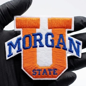 Op de afbeelding: Geborduurde patch met de tekst "MORGAN STATE" in blauw op een witte en oranje achtergrond. De patch heeft een unieke vorm met verlengde zijsecties. De patch wordt vastgehouden door een gehandschoende hand.