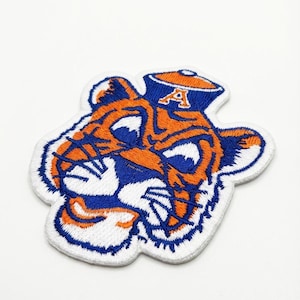 May include: Embroidered patch featuring a tiger head design in orange, blue, and white. The tiger is wearing a blue hat with the letter "A" on it. The patch has a white border and is likely for apparel or accessories.