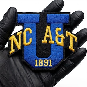 Könnte beinhalten: Gestickter Aufnäher mit den Buchstaben "NC A&T" in Gelb auf blauem und schwarzem Hintergrund. Das Jahr "1891" ist unten abgebildet. Der Aufnäher wird von einer behandschuhten Hand gehalten.