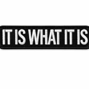May include: Black rectangular patch with rounded corners, embroidered with the phrase "IT IS WHAT IT IS" in white, bold, capital letters. The patch is designed to be sewn or ironed onto fabric.