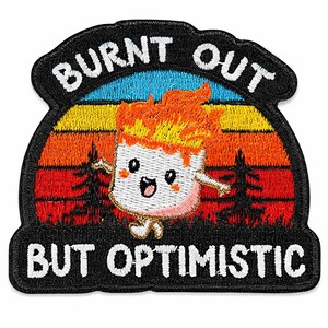 Könnte beinhalten: Gestickter Aufnäher mit einer Cartoon-Marshmallow-Figur vor einem Sonnenuntergangshintergrund. Das Marshmallow hat Flammen als Haare und lächelt. Der Text lautet "BURNT OUT BUT OPTIMISTIC" in weißen Buchstaben auf einem schwarzen Rand.
