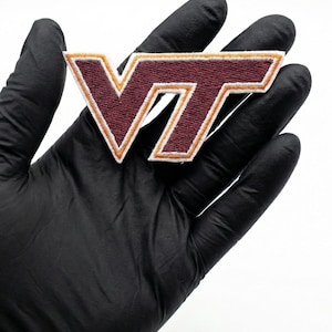 Könnte beinhalten: Gestickter Aufnäher mit den Buchstaben "VT" in Burgunderrot, goldener Umrandung und weißem Rand. Der Aufnäher wird vor einem schwarzen Hintergrund gehalten. Geeignet für Kleidung oder Accessoires.