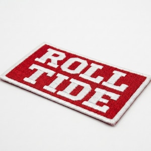 Könnte beinhalten: Gestickter rechteckiger Aufnäher mit den Worten "ROLL TIDE" in weißen Blockbuchstaben auf rotem Hintergrund. Der Aufnäher hat einen weißen Rand und ist leicht angewinkelt.