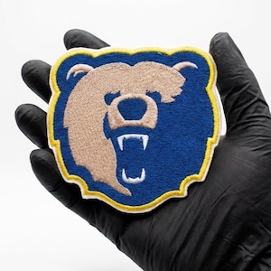 Op de afbeelding: Geborduurde patch met een berenkop met open mond en zichtbare tanden. De beer is bruin, tegen een blauwe achtergrond en geel omlijnd. De patch wordt vastgehouden door een gehandschoende hand.