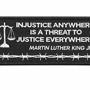 Puede incluir: Parche rectangular negro con texto bordado en blanco: "LA INJUSTICIA EN CUALQUIER LUGAR ES UNA AMENAZA PARA LA JUSTICIA EN TODAS PARTES" y "MARTIN LUTHER KING JR." Presenta una balanza de la justicia y alambre de púas.