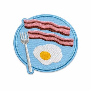 Può includere: Toppa ricamata raffigurante un piatto per la colazione con due fette di pancetta, un uovo fritto e una forchetta su sfondo azzurro. La pancetta è rosa con bordi marroni e l'uovo ha un tuorlo giallo.