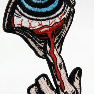 Könnte beinhalten: Gestickter Aufnäher mit einem detaillierten Auge mit blauer Iris, schwarzer Umrandung und roten Adern. Ein Finger mit rotem Streifen erstreckt sich vom Auge. Der Aufnäher hat einen schwarzen Rand auf weißem Hintergrund.