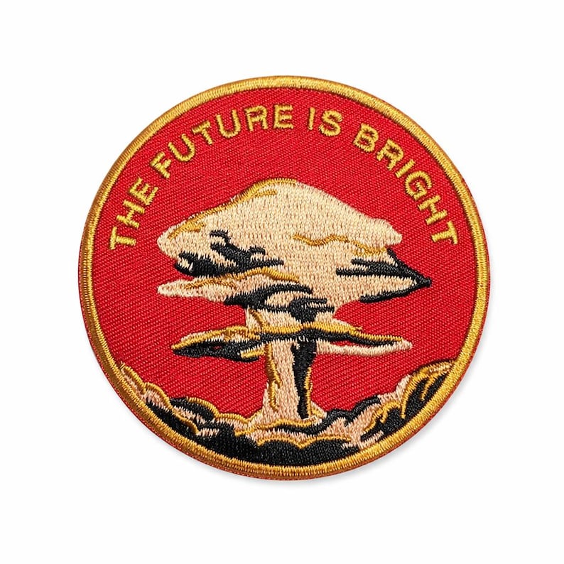 Peut inclure: &Eacute;cusson brod&eacute; circulaire avec un fond rouge et une bordure dor&eacute;e. Le motif repr&eacute;sente un champignon atomique en beige, noir et blanc. Le texte "THE FUTURE IS BRIGHT" est inscrit en arc de cercle sur le bord sup&eacute;rieur.