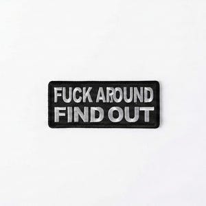 Può includere: Una toppa rettangolare nera con la frase "FUCK AROUND FIND OUT" ricamata in filo d'argento. La toppa è centrata su uno sfondo bianco.