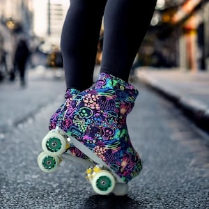 Könnte beinhalten: Rollschuhe mit einem farbenfrohen Schädel- und Neon-Design. Die Rollschuhe haben weiße Rahmen, grüne und gelbe Räder und stehen auf einer Asphaltfläche. Die Stiefel sind schwarz und mit einem lebendigen Muster aus Schädeln, Knochen und anderen Grafiken bedeckt.