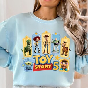Op de afbeelding: Een lichtblauw sweatshirt met ronde hals en een grafische print van Toy Story-personages zoals Rex, Jessie, Woody, Buzz Lightyear, Bullseye, een alien en Mr. Potato Head, met de tekst Toy Story 5 in het geel en rood.