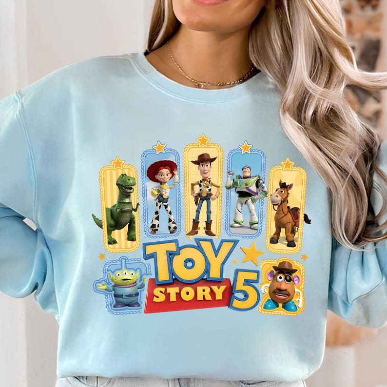 Op de afbeelding: Een lichtblauw sweatshirt met ronde hals en een grafische print van Toy Story-personages zoals Rex, Jessie, Woody, Buzz Lightyear, Bullseye, een alien en Mr. Potato Head, met de tekst Toy Story 5 in het geel en rood.