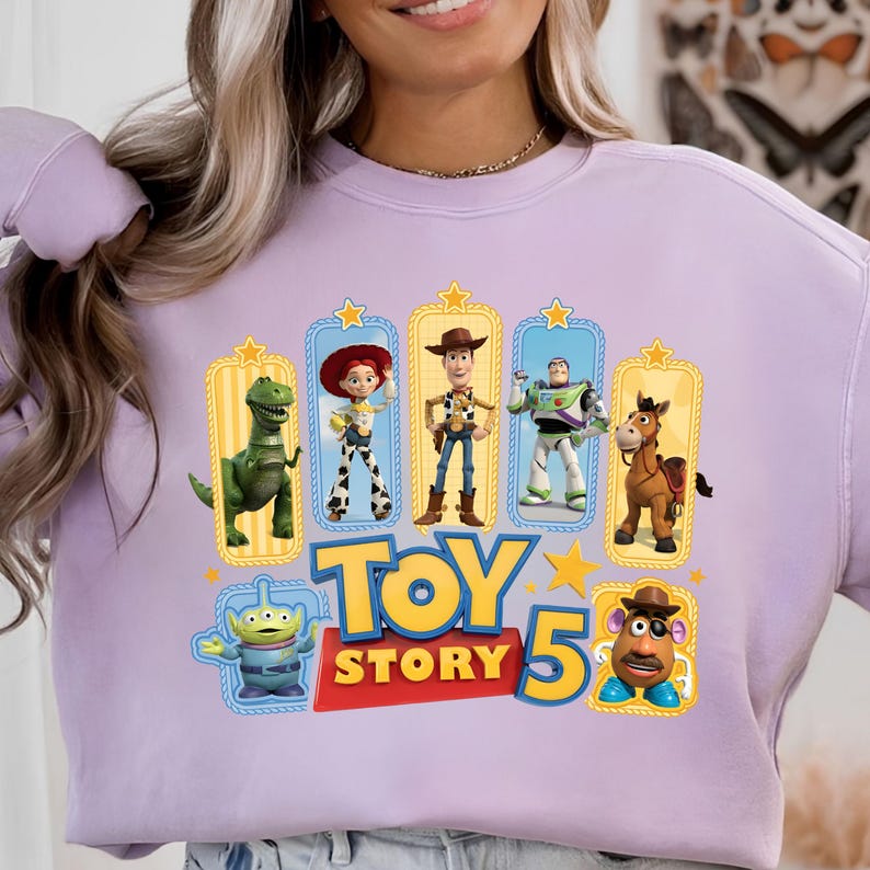 Op de afbeelding: Een lichtpaars sweatshirt met ronde hals en een grafische print van Toy Story-personages, waaronder Rex, Jessie, Woody, Buzz Lightyear, Bullseye, een alien en Mr. Potato Head, met de tekst Toy Story 5 in dikke gele en rode letters.