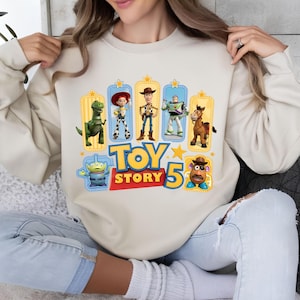 Op de afbeelding: Een beige sweater met ronde hals en een kleurrijke grafische print van Toy Story-personages, waaronder Rex, Jessie, Woody, Buzz Lightyear, Bullseye, een Alien en Mr. Potato Head, met de tekst Toy Story 5 in blauw en rood gedrukt.