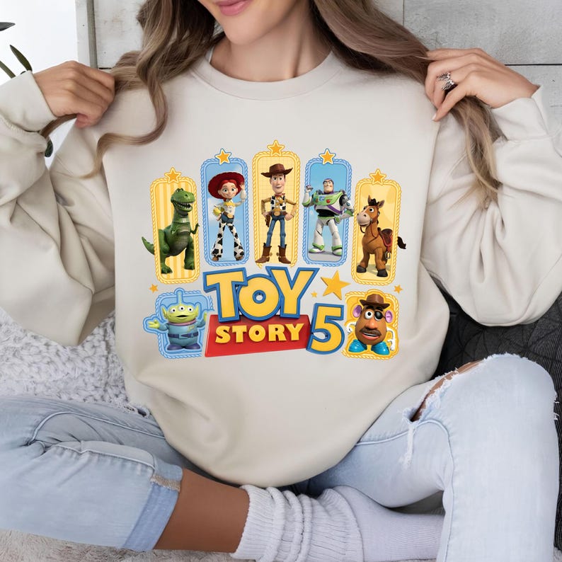 Op de afbeelding: Een beige sweater met ronde hals en een kleurrijke grafische print van Toy Story-personages, waaronder Rex, Jessie, Woody, Buzz Lightyear, Bullseye, een Alien en Mr. Potato Head, met de tekst Toy Story 5 in blauw en rood gedrukt.