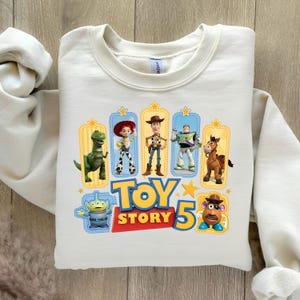 Op de afbeelding: Een cr&egrave;mekleurig sweatshirt met ronde hals en een kleurrijke grafische print van Toy Story-personages, waaronder Rex, Jessie, Woody, Buzz Lightyear, Bullseye, een alien en Mr. Potato Head, met de tekst Toy Story 5 in vetgedrukte letters.