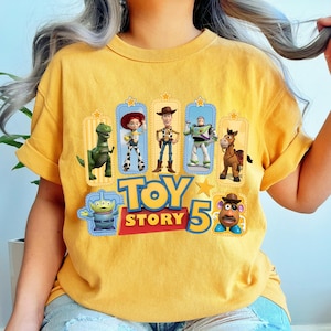 Op de afbeelding: Een mosterdgeel t-shirt met een grafische print van Toy Story-personages, waaronder Rex, Jessie, Woody, Buzz Lightyear, Bullseye, een alien en Mr. Potato Head, met de tekst "TOY STORY 5" in grote blauwe en rode letters.