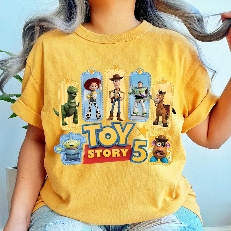 Op de afbeelding: Een mosterdgeel t-shirt met een grafische print van Toy Story-personages, waaronder Rex, Jessie, Woody, Buzz Lightyear, Bullseye, een alien en Mr. Potato Head, met de tekst "TOY STORY 5" in grote blauwe en rode letters.