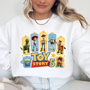 Op de afbeelding: Een wit sweatshirt met lange mouwen en een kleurrijke grafische print van Toy Story-personages, waaronder Rex, Jessie, Woody, Buzz Lightyear, Bullseye, een alien en Mr. Potato Head, met de tekst Toy Story 5 in het geel en rood.